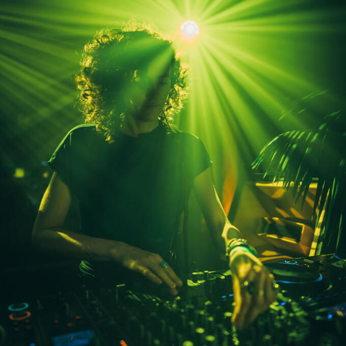 DJ Lamírla Live Behind the Decks DJ Lamírla performs behind the decks under bright green beams of light, focused on the mixer during her set.