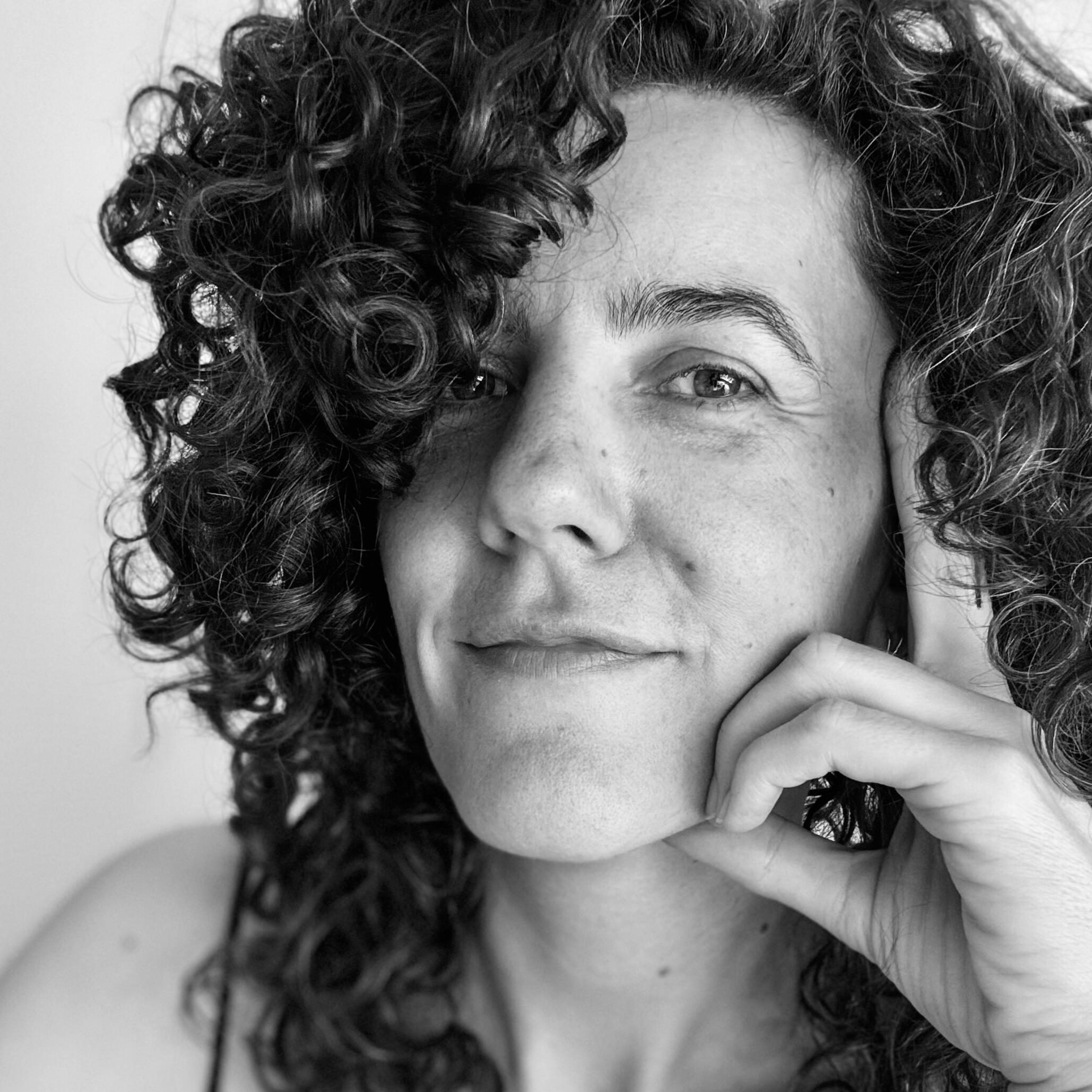 Black and White Portrait DJ Lamírla Black-and-white portrait of DJ Lamírla looking into the camera with a subtle smile, resting her chin on her hand. Her curly dark hair partly covers one side of her face.