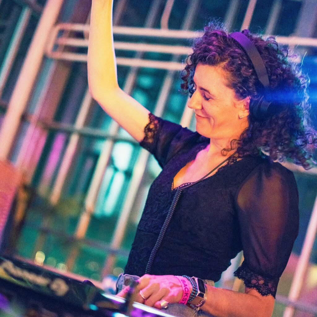 DJ Lamírla Live at Get in Synch DJ Lamírla performs behind the decks with one arm raised and a smile on her face. Colorful lighting creates a vibrant festival atmosphere.